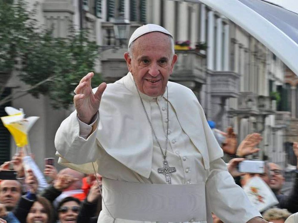 Pope Francis smiling with one arm outstretched.standing among a crowd of people, some taking photos and waving.
