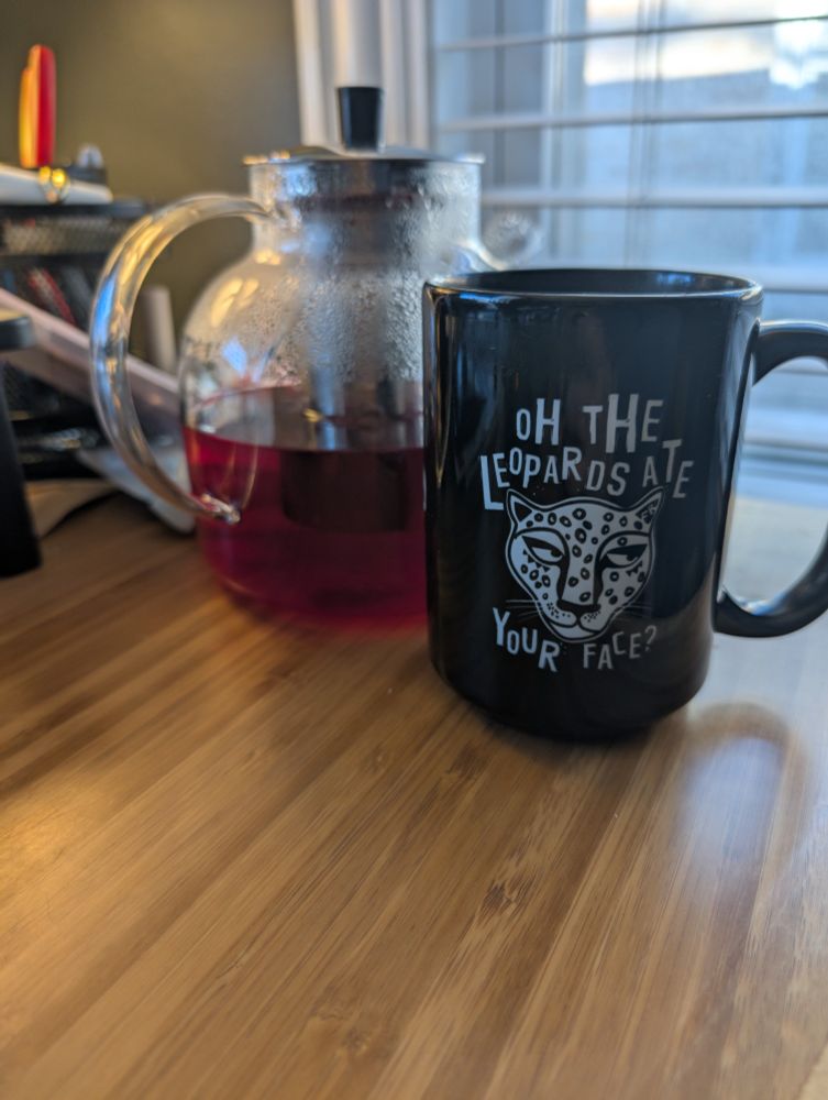 Steaming pot of tea with black mug that has the words " Oh the Leopards are your face?" along with the illustration of a stylized leopard rolling it's eyes. 