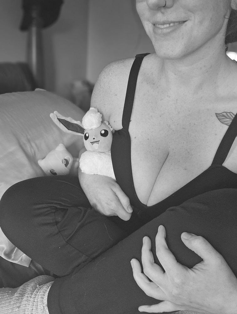Black and white photo of Lexy sitting cross-legged on bed, with stuffed Pokémon, in comfy pants and bra, smiling.