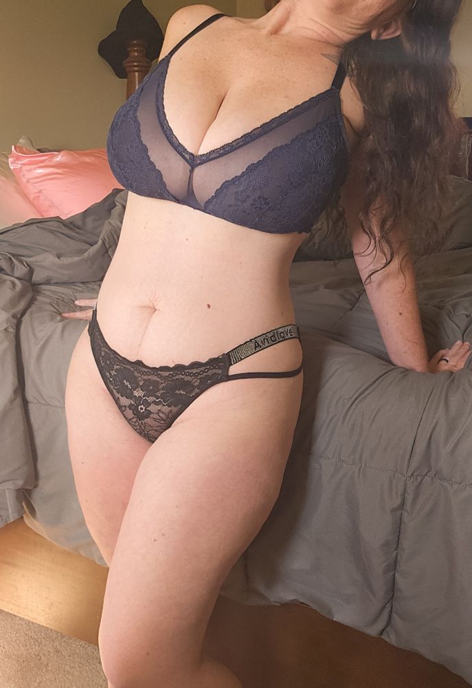 Lexy standing in a blue lace bralette and black lace panties next to a bed