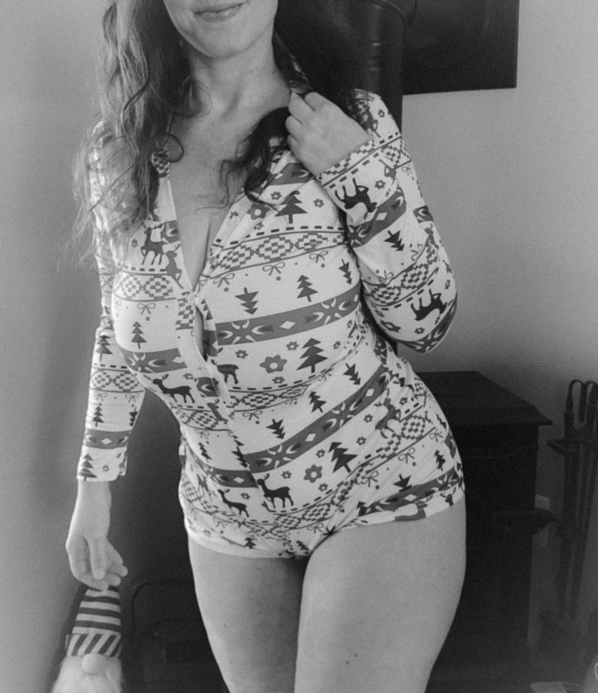 A black and white photo of a woman standing in front of a wood burning stove in a tightfit, shuper short, holiday themed onesie. Her cleavage about to bust through the seams while every curve is snugly being hugged.