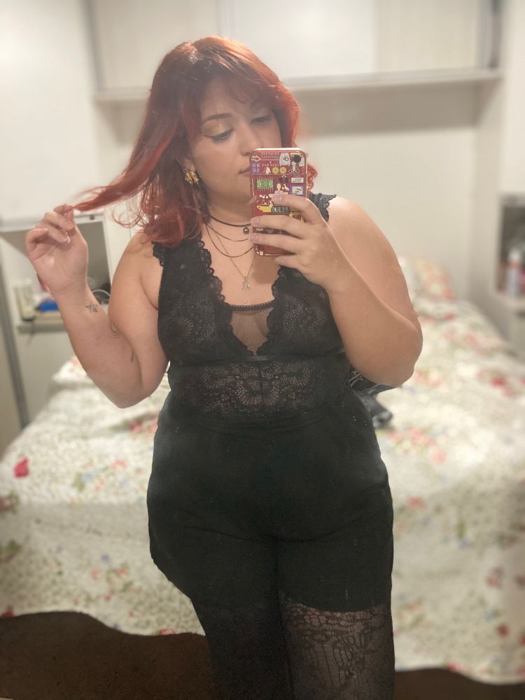 Foto de espelho minha: sou uma mulher de pele branca, cabelo ruivo avermelhado na altura dos ombros e estou vestindo um body de renda preta com um shorts preto e uma meia arrastão. 
