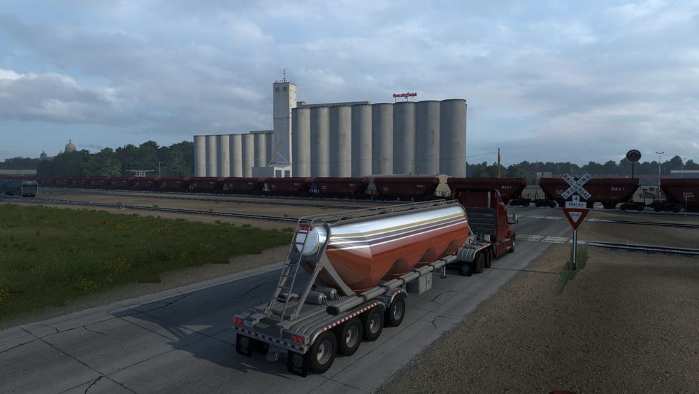 A picture from within American Truck Simulator(ATS) of my truck and trailer waiting at train tracks for a trim to go by, with Grain Silos in the background.