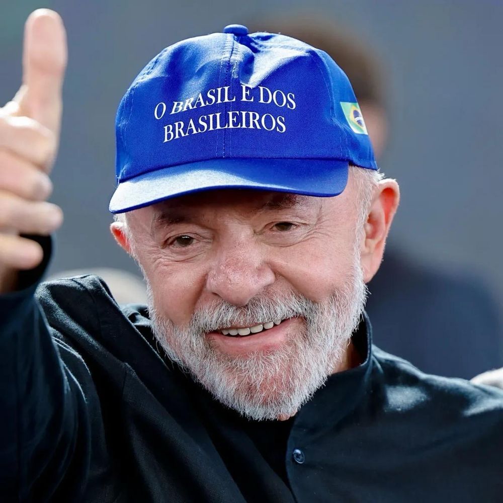 Foto do Presidente Lula sorrindo e levantando o braço para fazer um “jóinha” com a mão.

O Presidente usa um boné azul com o escrito “O Brasil é dos brasileiros” em branco na frente e uma pequena bandeira do brasil estampada na lateral.