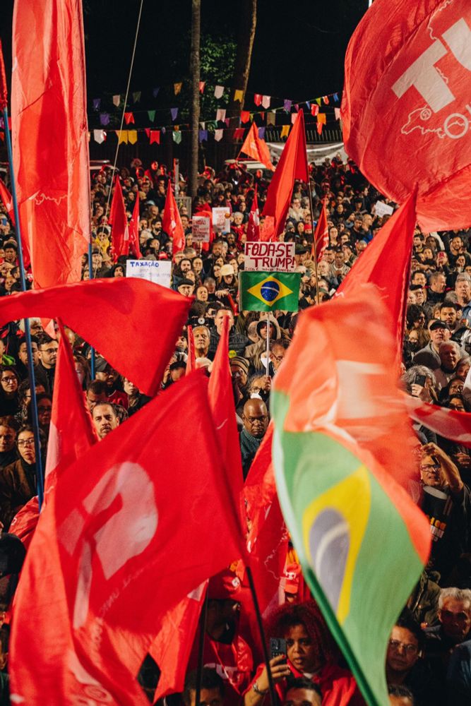 Foto de uma multidão em um protesto segurando grandes bandeiras de partidos, centrais sindicais e do Brasil.

No meio do povo, um homem mais velho segura uma placa caseira, onde há um papel com o escrito “Foda-se Trump! Essa é minha bandeira” e abaixo do papel uma pequena bandeira do Brasil.