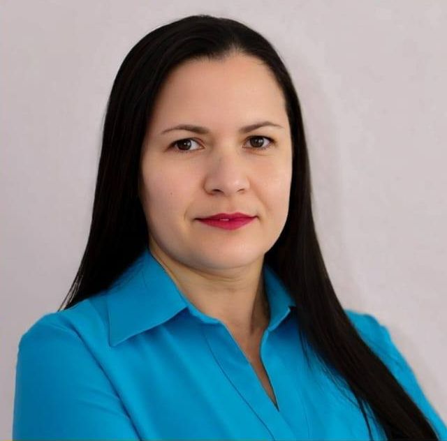 Foto frontal de Elisane Rodrigues dos Santos, mulher de pele clara, de cabelos pretos e longos, olhos castanhos, olhando para a câmera com um pequeno sorriso. Ela veste uma camisa azul clara.