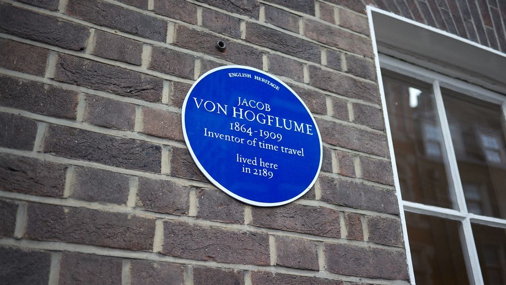 Looking up at brick wall next to a window, on which is hanging a circular Blue Plaque. It used to be in Golden Square, Soho. The text on it says "English Heritage. Jacob Von Hogflume. 1864-1909 Inventor of time travel. Lived here in 2189."
