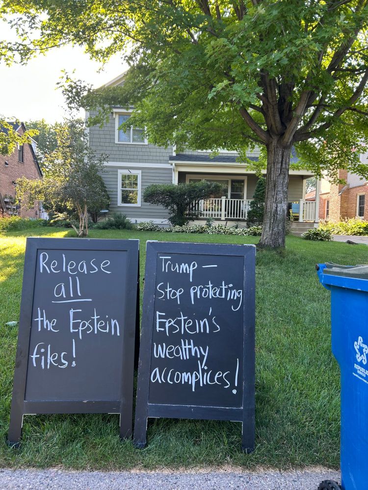 Lawn sign reading "Trump — stop protecting Epstein's wealthy accomplices! Release all the Epstein files!' with trash bin in background.