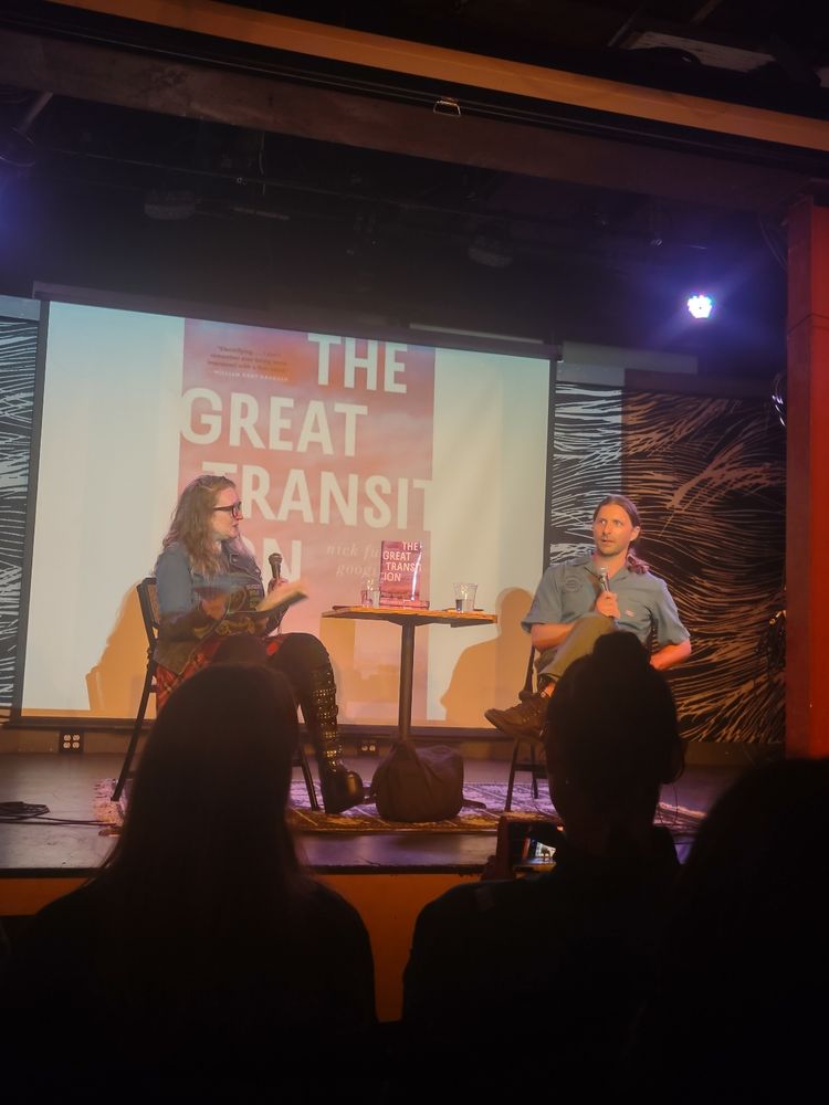 Author Nick Fuller Googins and interviewer Liberty Hardy sit on stage with microphones discussing Nick's book The Great Transition, which is projected on a screen behind them. 