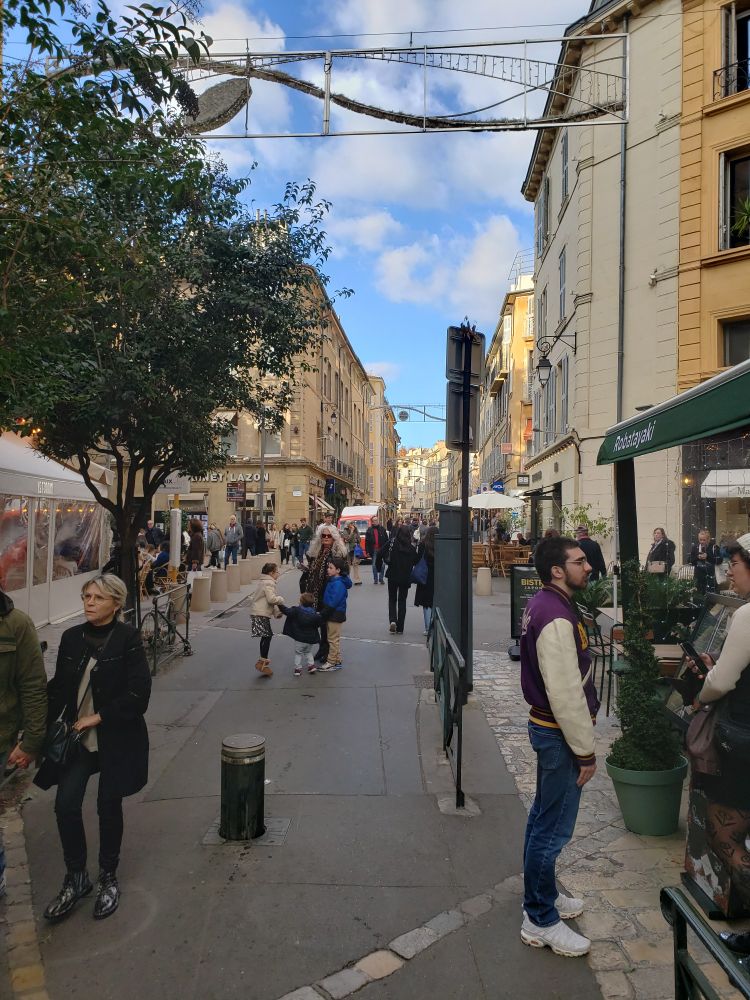 Lively street scene, all pedestrians except for a small six-seat electric bus in the mid-ground, with four-story apartment buildings on either side and small shops and cafés on the ground floor. People stopping to chat. Control bollard in foreground can be lowered to allow access for delivery vehicles and public mini-buses. Rue D'Italie, Aix-en-Provence.