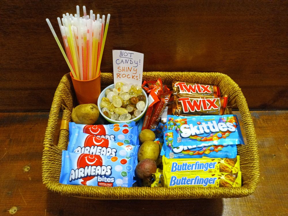 A basket containing an inviting array of treats: full-sized Butterfinger, Twix, Skittles, and Airheads candies; a cup full of glow sticks; a bowl of tumbled quartz crystals marked NOT CANDY! SHINY ROCKS!; and several potatoes.