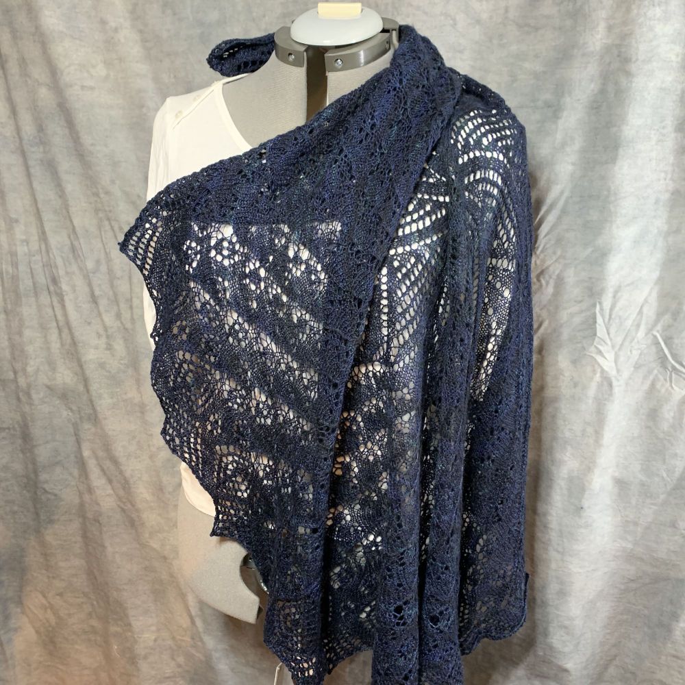 A lace shawl of mostly blue but with teal highlights on a dress form with a wrinkly grey backdrop