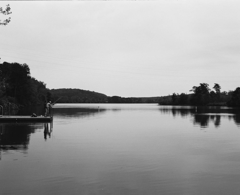 A black and white photograph of a calm lake with reflections of the sky. Trees can be seen in the distance and there is a man fishing from a docking the left. 