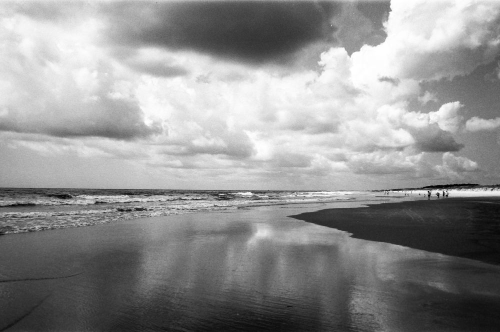 A black and white photograph of the beach with reflections of the cloudy sky on a sheen of water on the beach. There are large white and grey clouds overhead. 
