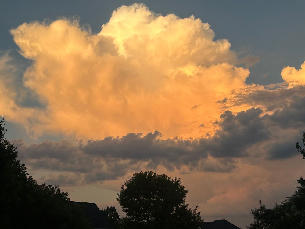 Large puffy clouds at sunset, alternating between orange and yellow and gray above the line of trees and houses.
