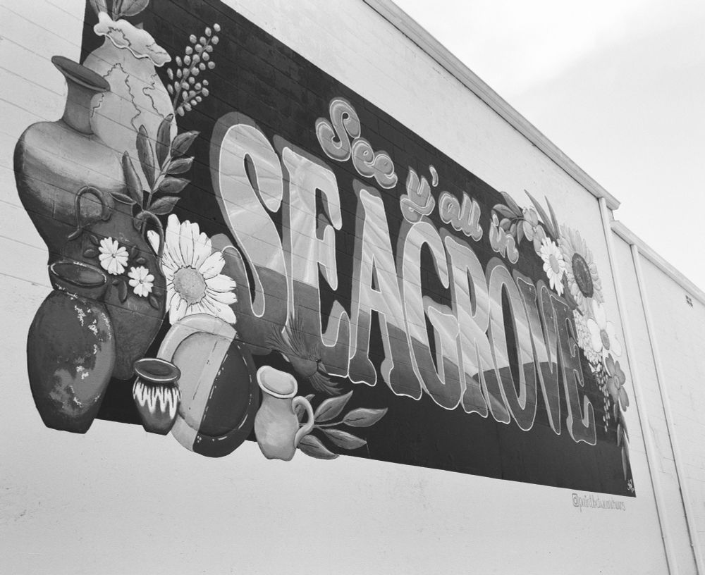 A black and white photograph of a mural painted  the side of a building that read “See y’all in Seagrove”. Flowers and pottery adorn the mural. 