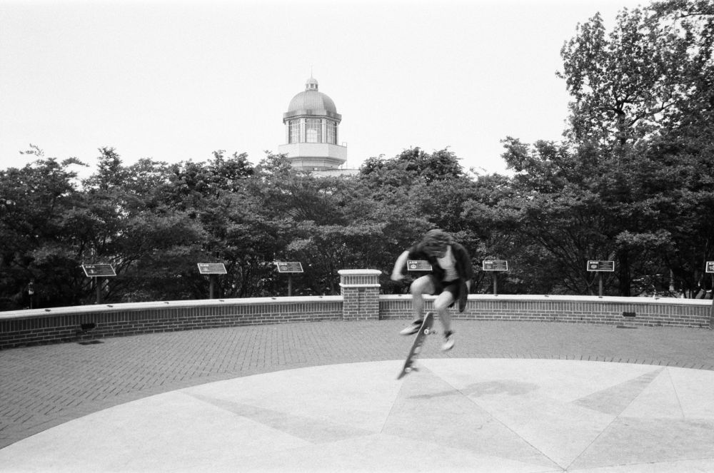 Black and white photograph of a skateboarder doing a flip with his board on a circular plaza on a college campus. There is a domed building in the background. 