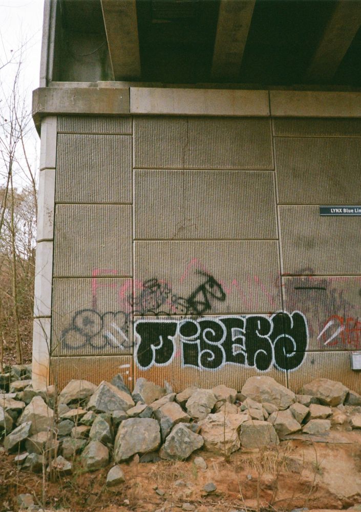Graffiti on a concrete support wall of an underpass with stones at the base and brown grass and weeds. 