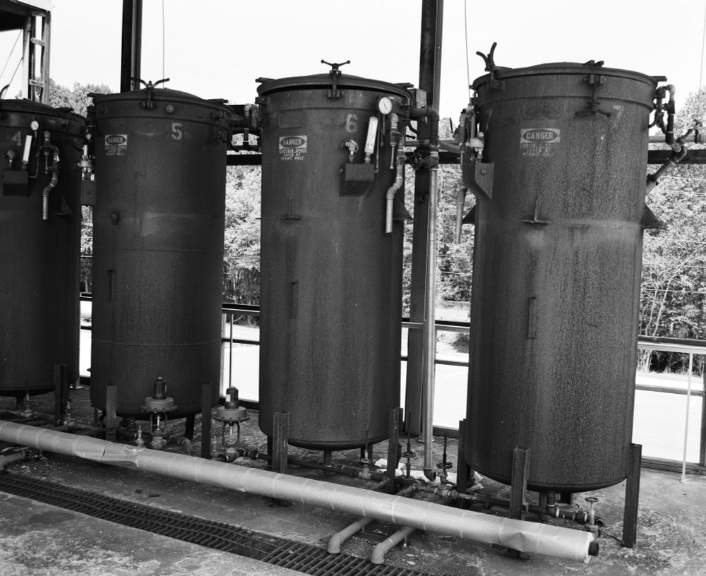 Four tall, industrial metal tanks are lined up under an awning behind a building. Each has gauges and pipes attached. 