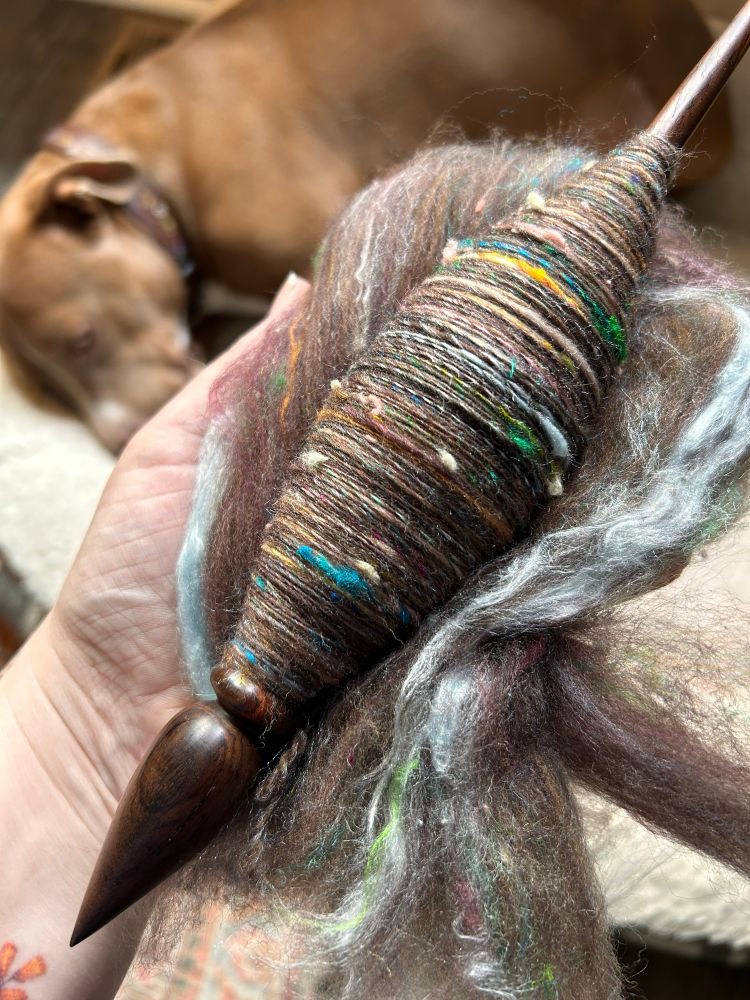 A Russian spindle from Willow’s Spindles with some dark brown fiber spun around it. The fiber has bits of blue, green, yellow, and pink running through it. 