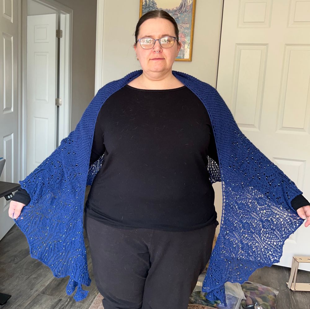 A woman in all black faces the camera with a lace and beaded bright blue shawl over her shoulders. The shawl is a crescent shape. 