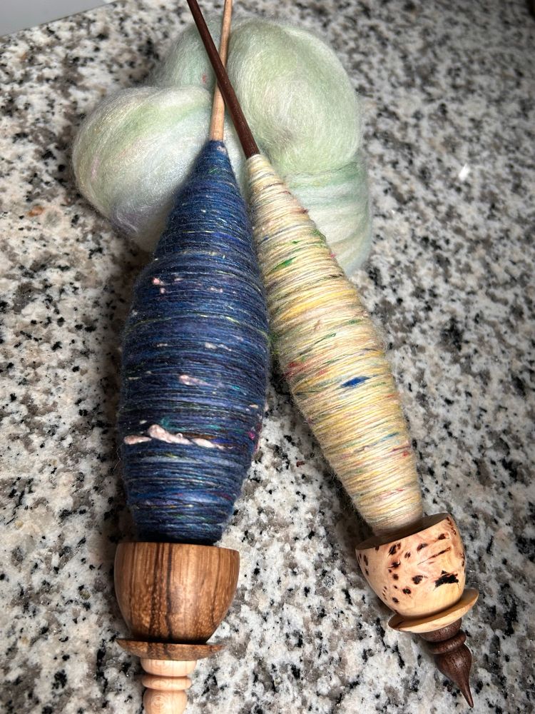 Two Woodland Handcrafts tea cup support spindles rest on a coil of light green fiber. The spindle on the left has a dark blue cop on it and has a zebrawood whorl. The spindle on the left has a bright yellow cop and its whorl is carved from a burl. 