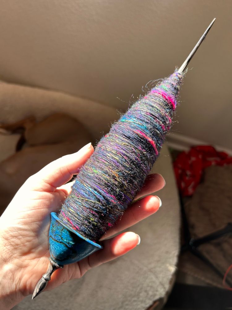 A hand holds a Spanish Peacock Tibetan spindle with a blue whorl. It has a large cop of black, blue, pink, yellow, and orange fiber. 