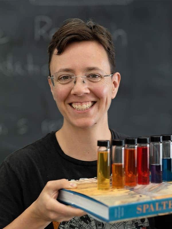 Scientist/artist standing in front of a blackboard with a book and a big smile, balancing glass tubes with multiple colors inside.