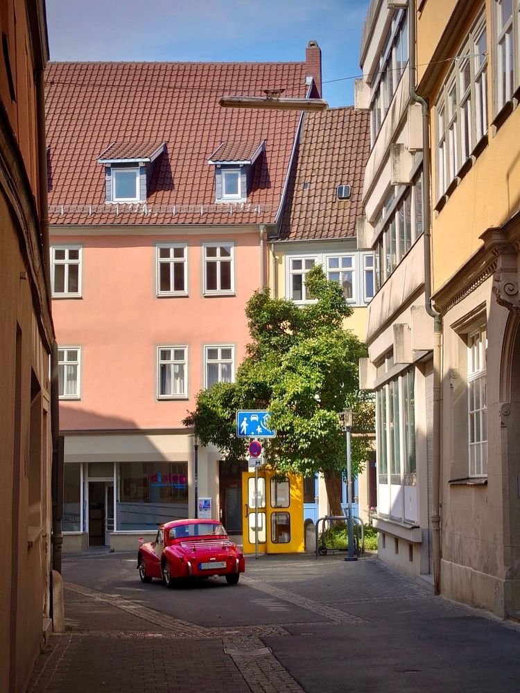 Blick in eine Altstadtgasse: Ein kleiner roter Oldtimer fährt an einem gelben Telefonhäuschen vorbei. Die Tür ist offen, es steht unter einem Baum, darüber ist ein blaues Schild „Spielstraße“ zu sehen.