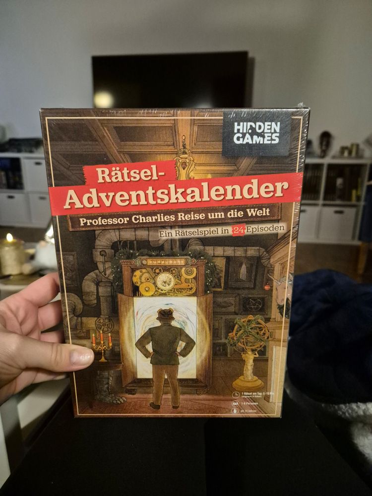 Hand hält einen Rätsel-Adventskalender von Hidden Games mit dem Titel Professor Charlies Reise um die Welt.