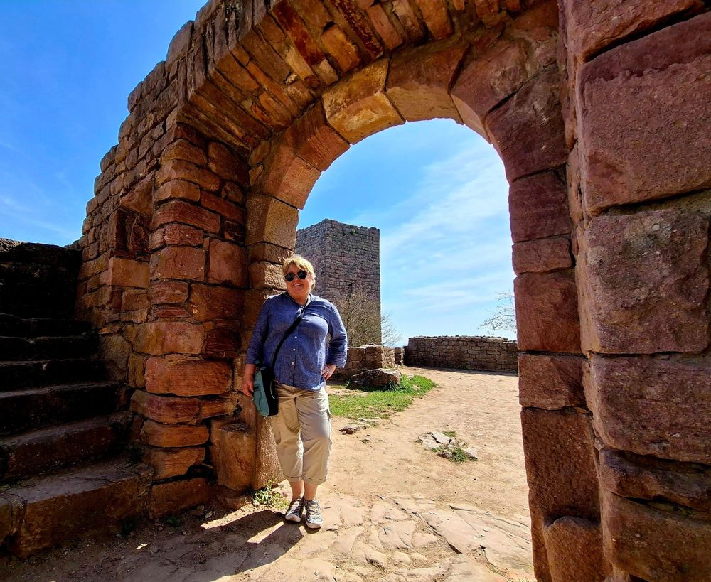 A smiling person stands beneath a stone archway of an old castle or fortification made from reddish-brown stones. They are wearing sunglasses, a blue shirt, beige capri pants, and sneakers, with a small bag slung over the shoulder. Behind them, the path leads to a partially visible stone tower and low stone walls. The sky is clear and blue, and the surrounding area is open and sunlit, evoking a warm, historical atmosphere.