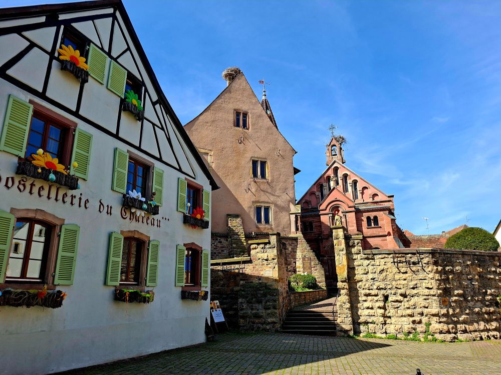 The image shows a charming European village scene with traditional Alsatian architecture. On the left, there's a half-timbered building with green shutters and colorful flower decorations on the window boxes, labeled "Hostellerie du Château." In the center, a narrow, older stone building with a steep roof stands, topped with a large stork's nest. To the right, a small red stone church or chapel with arched windows and a statue above the entrance is visible. The foreground is paved with cobblestones, and a stone stairway leads up to the chapel. The sky is clear and blue, enhancing the picturesque, peaceful atmosphere.