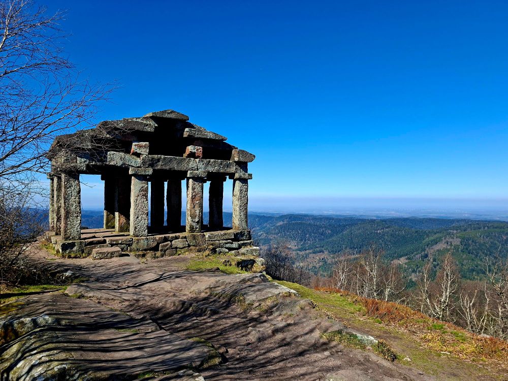 A striking stone pavilion with a weathered, ancient appearance stands on the edge of a rocky outcrop overlooking a vast, forested valley. The structure, made of large stone pillars and slabs, resembles a classical temple ruin. Bare trees frame the left side of the image, while the right opens up to a sweeping view of rolling green hills and distant plains under a clear, vibrant blue sky. The bright sunlight enhances the textures of the stone and the natural landscape, creating a dramatic and serene scene.