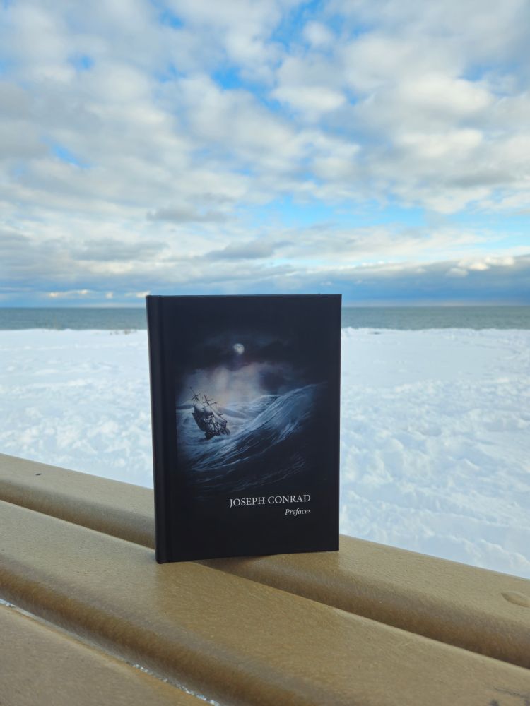  image features Lake Ontario in the far background, with rolling billowy clouds and a blue sky above and piles of snow on the beach. The book, JOSEPH CONRAD PREFACES, is featured in the foreground atop a brown wood railing. It has a black/navy cover with an image of a tall sailboat on tumultuous waves and a darkened sky with a sliver of moon visible.
