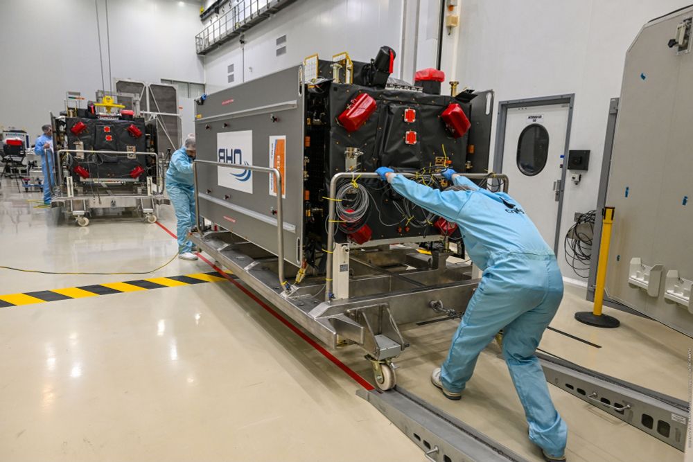 All'interno di una camera pulita, due persone, che indossano tute azzurre da camera bianca, scaricano il satellite Galileo. Ciascuna persona è posizionata a una delle estremità del satellite. 