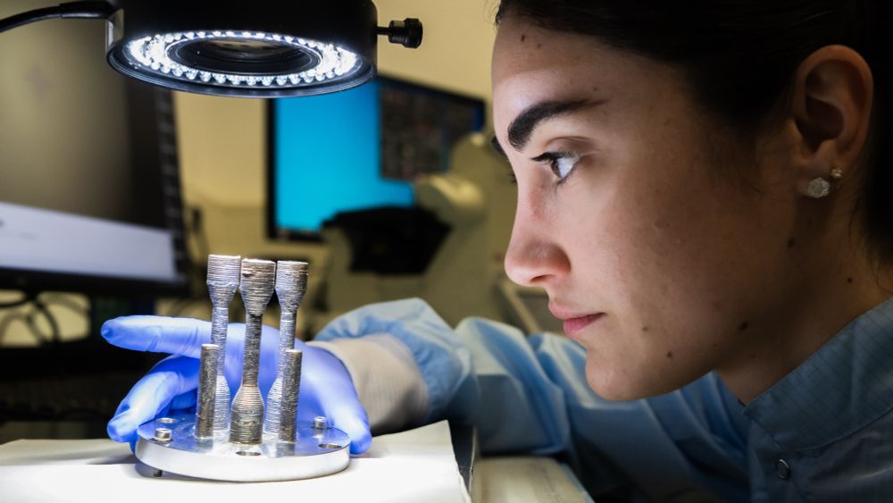 Ispezione del primo componente metallico mai stampato in 3D nello spazio. Un'operatrice, in primo piano di profilo a destra nell'immagine, ispeziona dei componenti metallici montati su un piccolo supporto tondo, sotto una lampada di laboratorio. La scienziata indossa camice e guanti da lavoro.