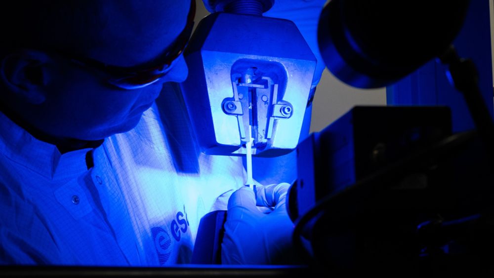 Ispezione del primo componente metallico mai stampato in 3D nello spazio. La foto è illuminata di una delicata luce blu. Sulla sinistra, con indosso un camice con il logo ESA e occhiali di protezione, un tecnico ispeziona il componente, al centro in primo piano, stampato in 3D nello spazio. 