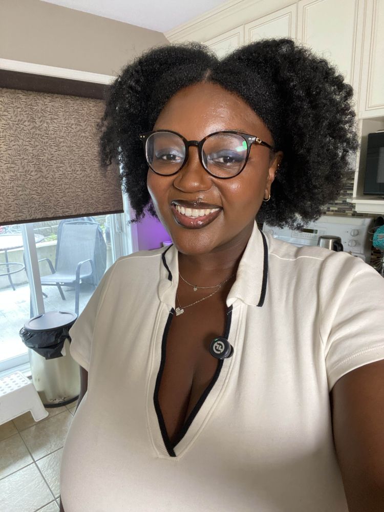 Selfie of Pink. She is standing in a kitchen. She has deep brown skin, afro hair styled in a braid out. She is wearing a vneck white polo shirt.