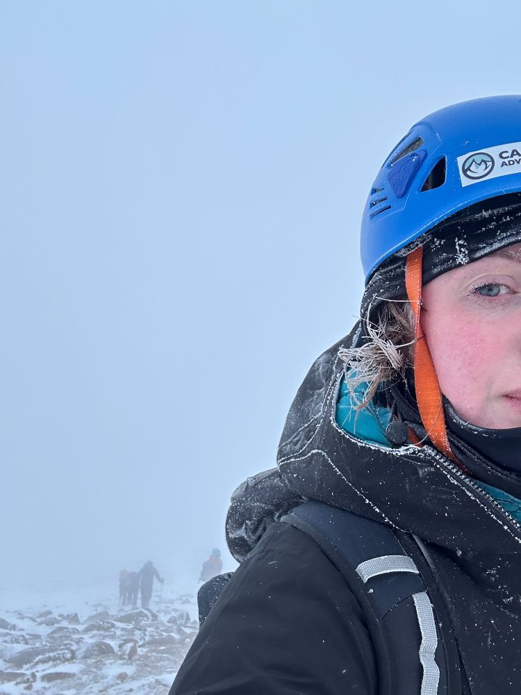 A women standing amongst a whiteout on a mountain wearing a blue helmet and hiking gear, hair strands and eyelashes covered in ice, and a silhouette of a group of hikers behind her.