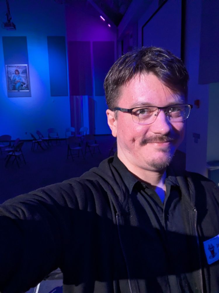 Selfie with stage light hitting DJ wearing glasses, black hoodie and a buttoned black shirt looking toward camera with the Space Center Complex stages and chairs covered by blue lighting in the background.
