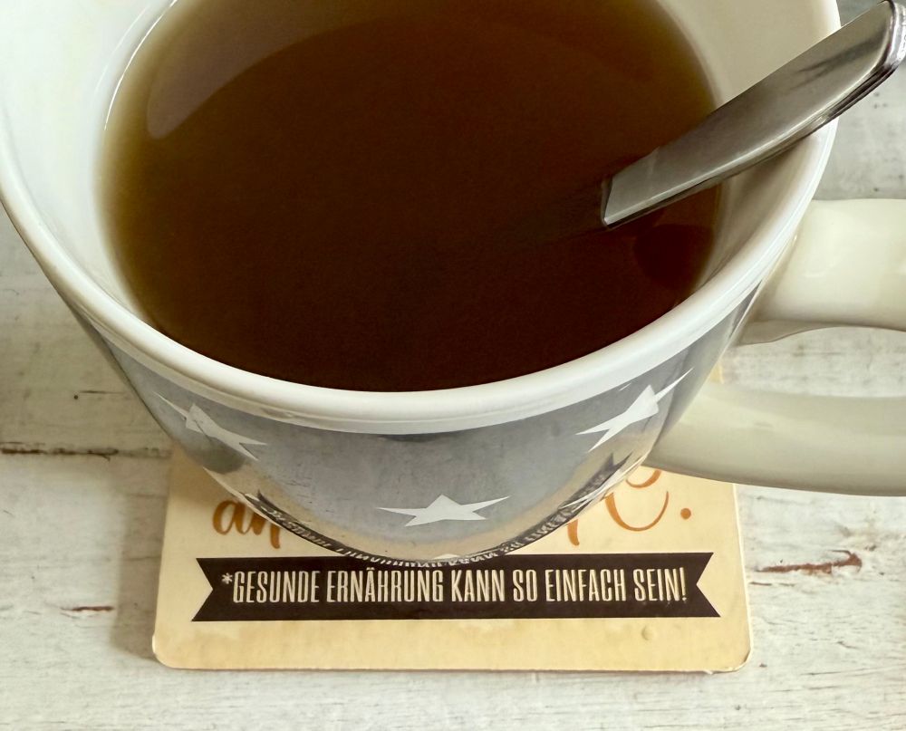 Halber Blick (von oben) in eine Tasse Tee, die auf einem Tischuntersetzer steht. Darauf zu lesen „Gesunde Ernährung kann so einfach sein“.