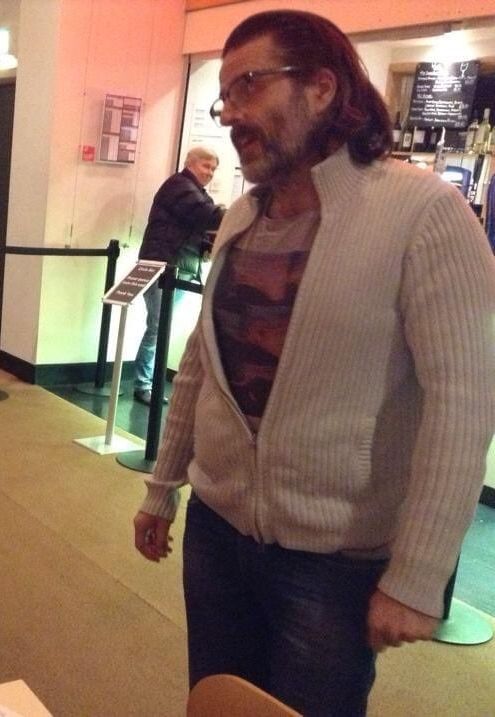 Con O'Neill with longer hair and glasses. wearing a light hoodie with zipper and blue jeans, he has his arms by his side. He has a beard.