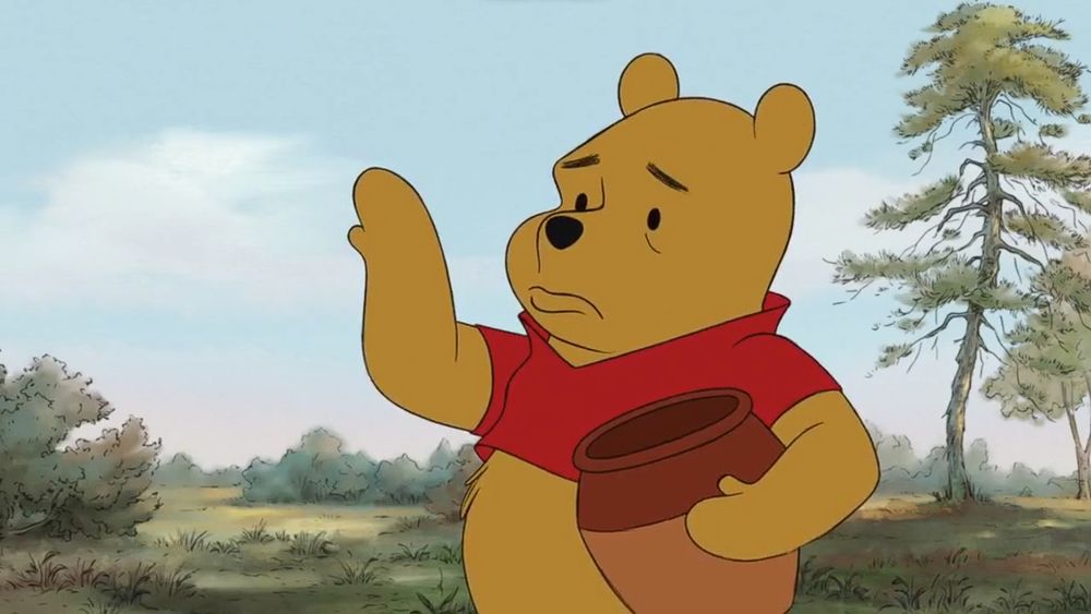 A screenshot from Winnie the Pooh 2011, it's Poohbear looking at his hand in sadness as his other hand is holding an empty pot of honey
The background is a forest in a sunny day