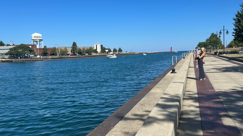 Lake Michigan lakefront in Kenosha, WI showing walking path with blue sky and lighthouse in background, as well as a white Kenosha, WI water tower and some people enjoying the sunny day on the path by the water