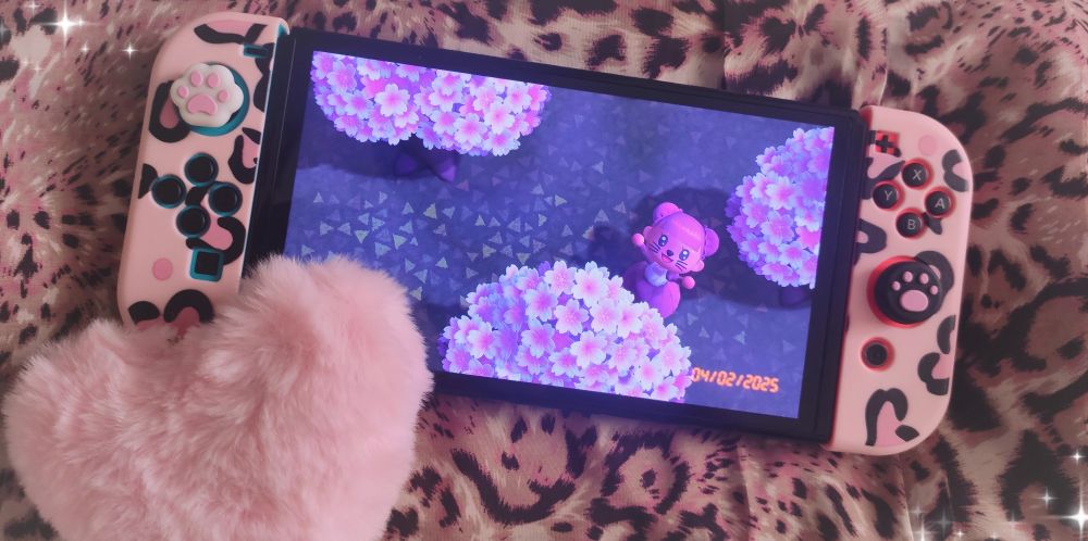 Pink and patterned image of SplingleSploink in animal crossing, waving up at the camera surrounded by cherry blossom trees. Their switch of course has pawpad joycons! A fluffy pink heart is layered on top of the switch and sparkles frame the corners of the image.
