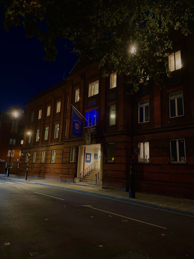 A brick building at night with its entrance and several windows lit from inside. One upper window glows blue. The street outside is quiet and empty, with a few streetlights and tree branches visible above.