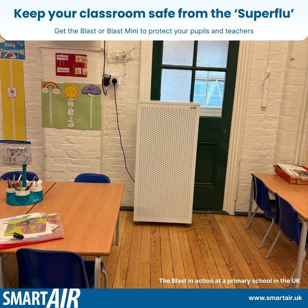 A classroom setting in a primary school with wooden flooring and white brick walls. Two tables with blue chairs are arranged on the left and right sides, with stationery and books placed on top. A large white air purifier (Blast by Smart Air), stands against the wall near a green door. Colourful educational posters are displayed on the wall. Text at the top reads: “Keep your classroom safe from the ‘Superflu’ – Get the Blast or Blast Mini to protect your pupils and teachers.” Branding at the bottom shows “SMARTAIR” and the website “www.smartair.uk.”