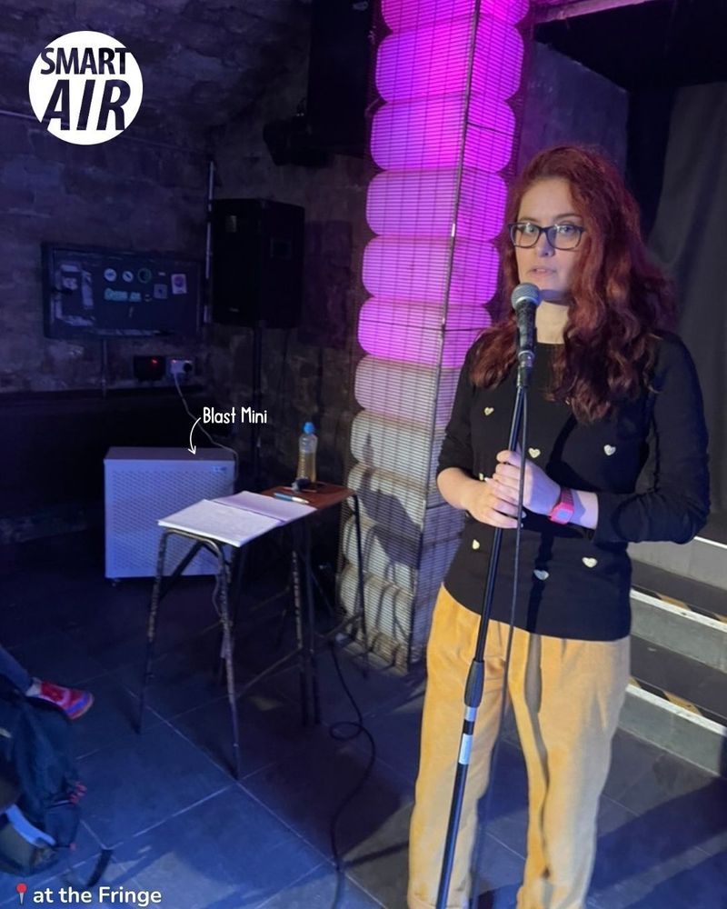 The Blast Mini air purifier placed beside a performer on stage at the Fringe festival. The performer, with long wavy hair, wears a black shirt with heart patterns and mustard yellow pants, standing in front of a microphone. The venue features a stone wall, pink lighting, and a small table with a water bottle.