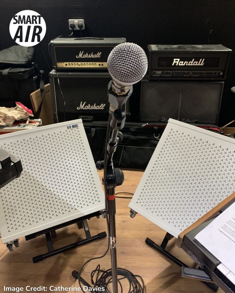 A microphone on a stand in front of two Smart Air Blast Mini purifiers in a rehearsal room, with Marshall and Randall amplifiers in the background.
