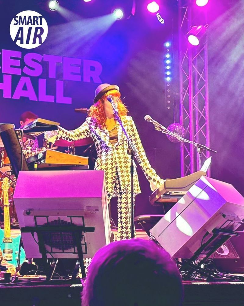 The Anchoress performing on stage under purple lighting with Smart Air Blast Mini purifiers placed near the instruments for clean air during the concert.